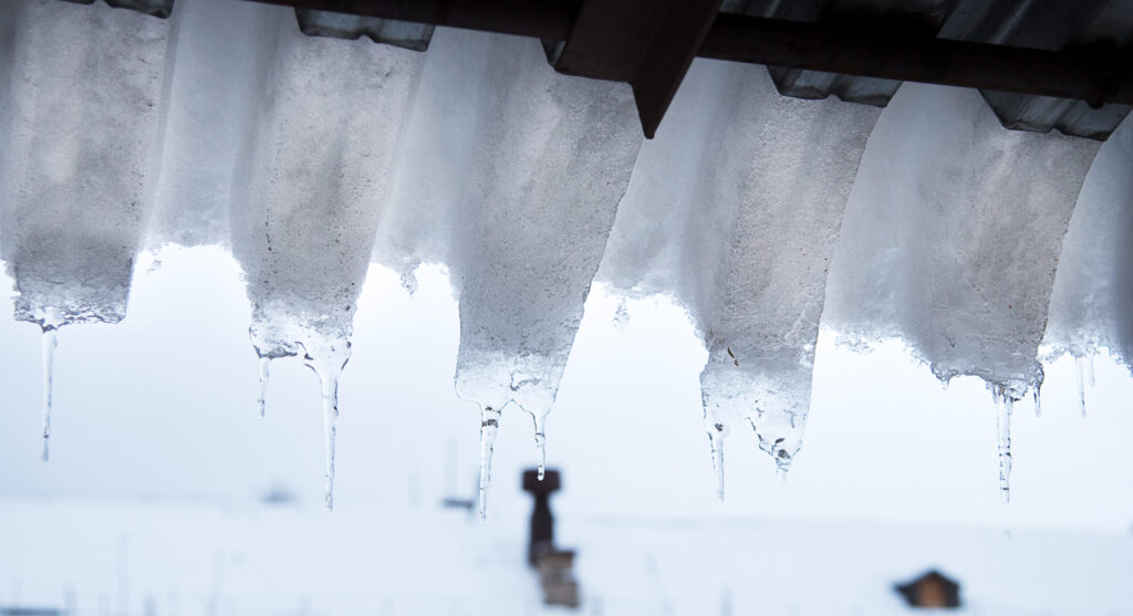 Icicles hanging from the edge of a snow-covered roof hint at the presence of ice dams, with a blurry snowy landscape and a few indistinct structures visible in the background.