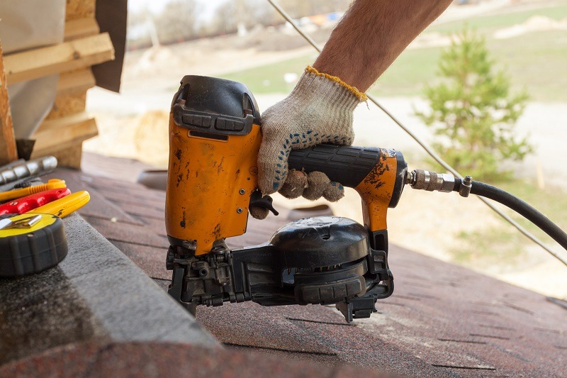 A person wearing a work glove uses a yellow pneumatic nail gun to secure roofing shingles on a sloped roof, with tools and a blurred outdoor background visible.