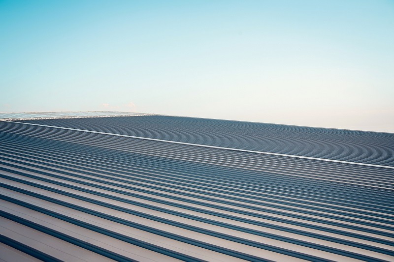 A wide view of a modern metal roof with parallel ridges and lines, under a clear blue sky. The roof’s surface appears sleek and reflective, extending into the distance.