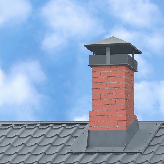 A red brick chimney with a metal cap rises from a gray, tiled rooftop against a blue sky with scattered white clouds.