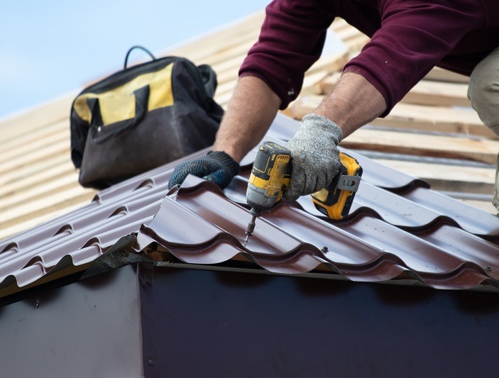 A person wearing gloves uses a power drill to secure a sheet of metal roofing onto a wooden roof frame. A tool bag is nearby on the roof.