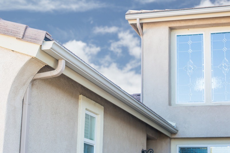 Close-up of a beige stucco house showing white rain gutters along the roof edge, decorative glass windows, and a blue sky with scattered clouds in the background.