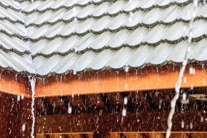 Heavy rain pours down on a sloped, tiled roof with water flowing off the edges. The roof sits above wooden beams, and large raindrops are visible in the foreground.