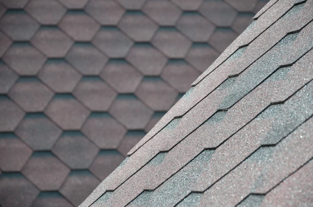 Close-up view of brown asphalt roof shingles, arranged in a hexagonal pattern. The image shows part of a rooftop with a slanted section intersecting a flat section, highlighting the texture and pattern of the shingles.