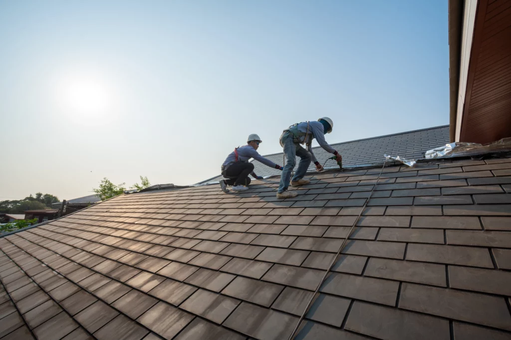 Two workers wearing safety harnesses and helmets are repairing or installing shingles on a sloped house roof under bright sunlight. The sky is clear and the sun is low on the horizon.