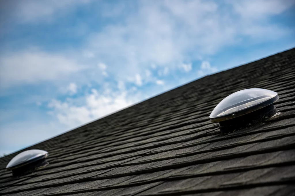 Close-up of a dark shingled roof with two dome-shaped skylights under a partly cloudy blue sky.