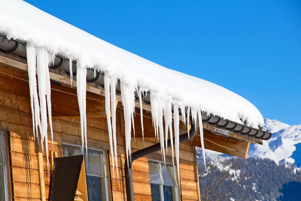 A wooden house with a snow-covered roof and long icicles hanging from the eaves, set against a clear blue sky and distant snow-capped mountains.