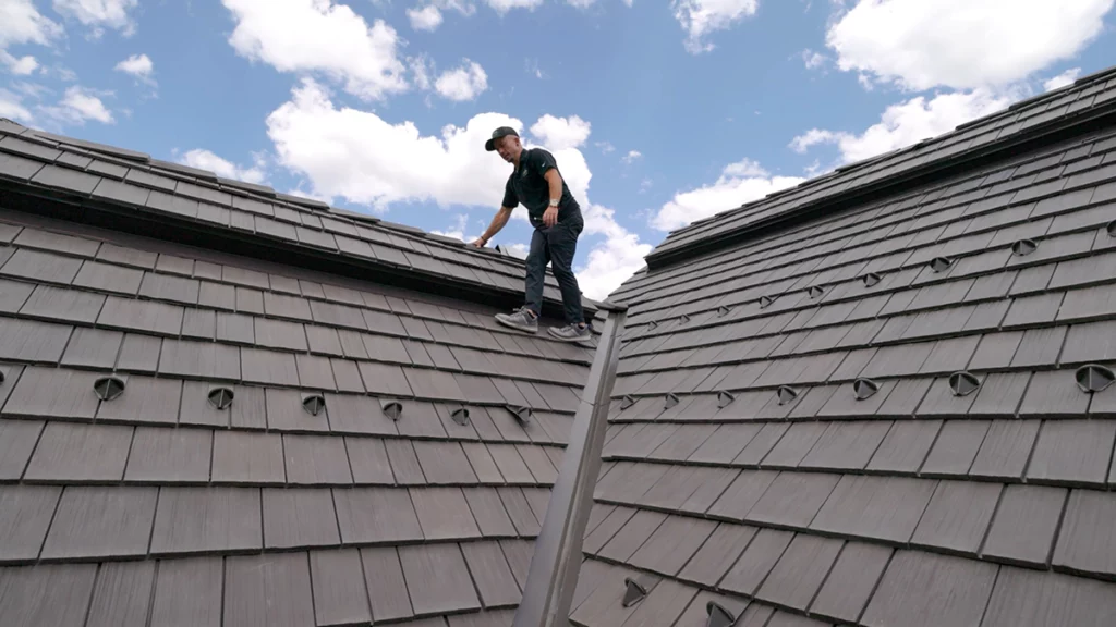 A person wearing a cap and dark clothing stands on a steep, gray shingled roof under a partly cloudy sky, looking down at the roof surface while holding onto the ridge.