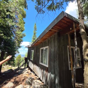 A rustic wooden cabin with white-framed windows situated on a rocky, tree-lined hillside under a blue sky.