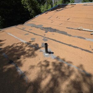 A flat roof covered in worn, tan mineral-surfaced roofing felt with a single metal pipe protruding through a sealed flashing.