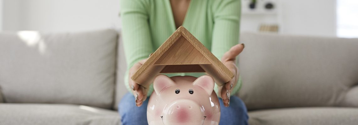 Happy woman who plans buying house or taking mortgage holding roof above piggy bank